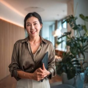 Positioned within a modern corporate environment, a mid-aged Asian businesswoman of striking beauty stands in her office, poised with a digital tablet, using it to execute digital tools and wireless technology to perfection in her strategic endeavors.